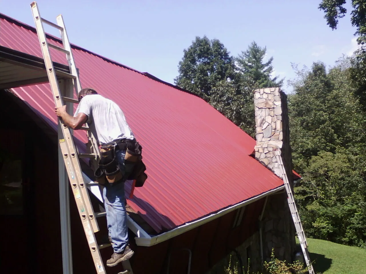 Roofer on a ladder beside a red metal roof and stone chimney — Soffit & Fascia Repair by Roofing Brothers in Mission Hill